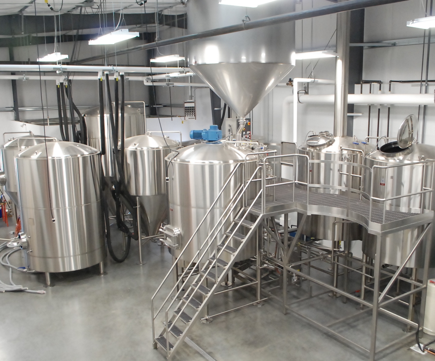 A room filled with different sized brewery fermentation and storage tanks with a ladder leading up to a platform to view inside the tanks.